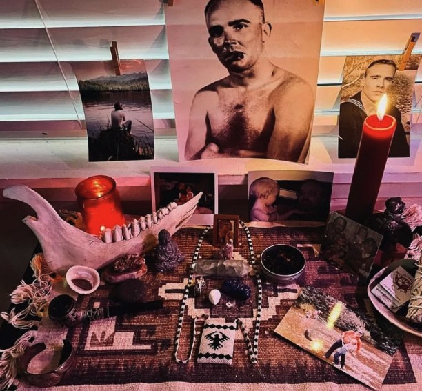 A memorial altar with photographs, candles, sage, a jawbone, and keepsakes arranged on a woven cloth