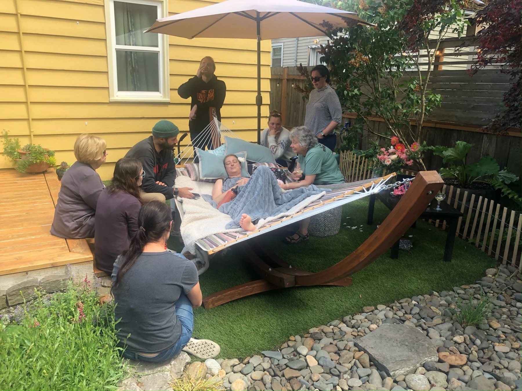 Friends and family gather around a loved one resting in a hammock in a sunny backyard garden