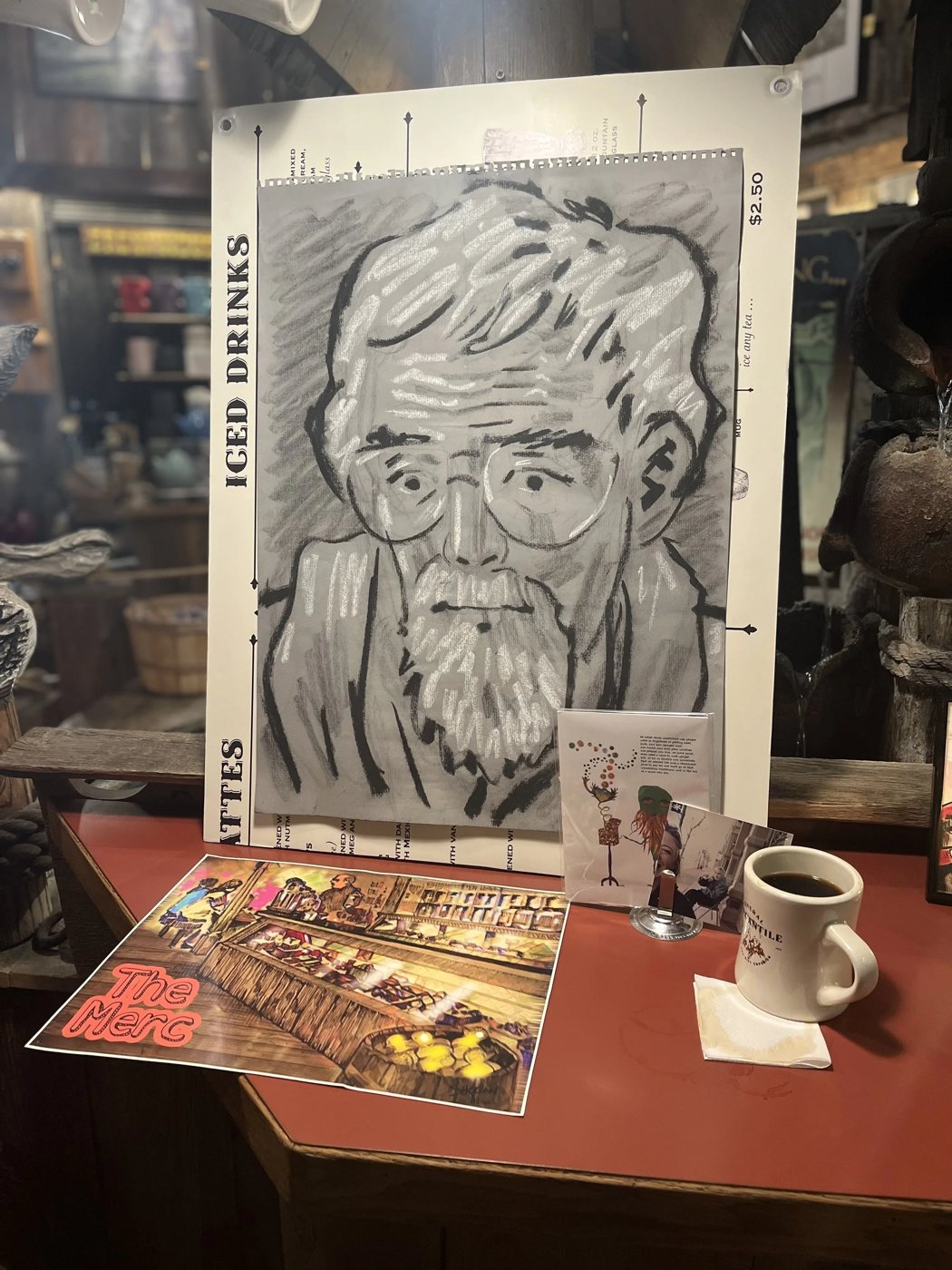 A charcoal portrait of a bearded man in glasses displayed with a cup of coffee and keepsakes at The Merc